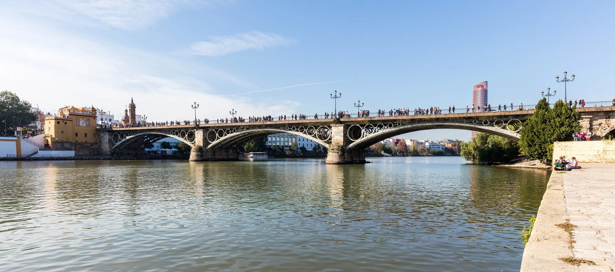 The iron Isabel II bridge crossing the Guadalquivir in Seville, with Triana's yellow chapel and towers on the left bank and people walking across