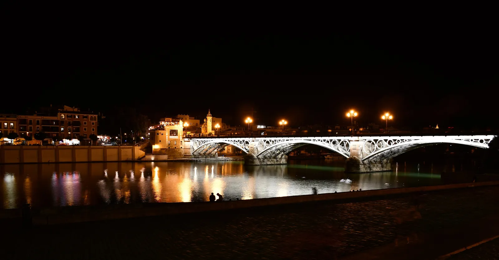 The lit Isabel II bridge reflected in the river at night, with lights along the bank