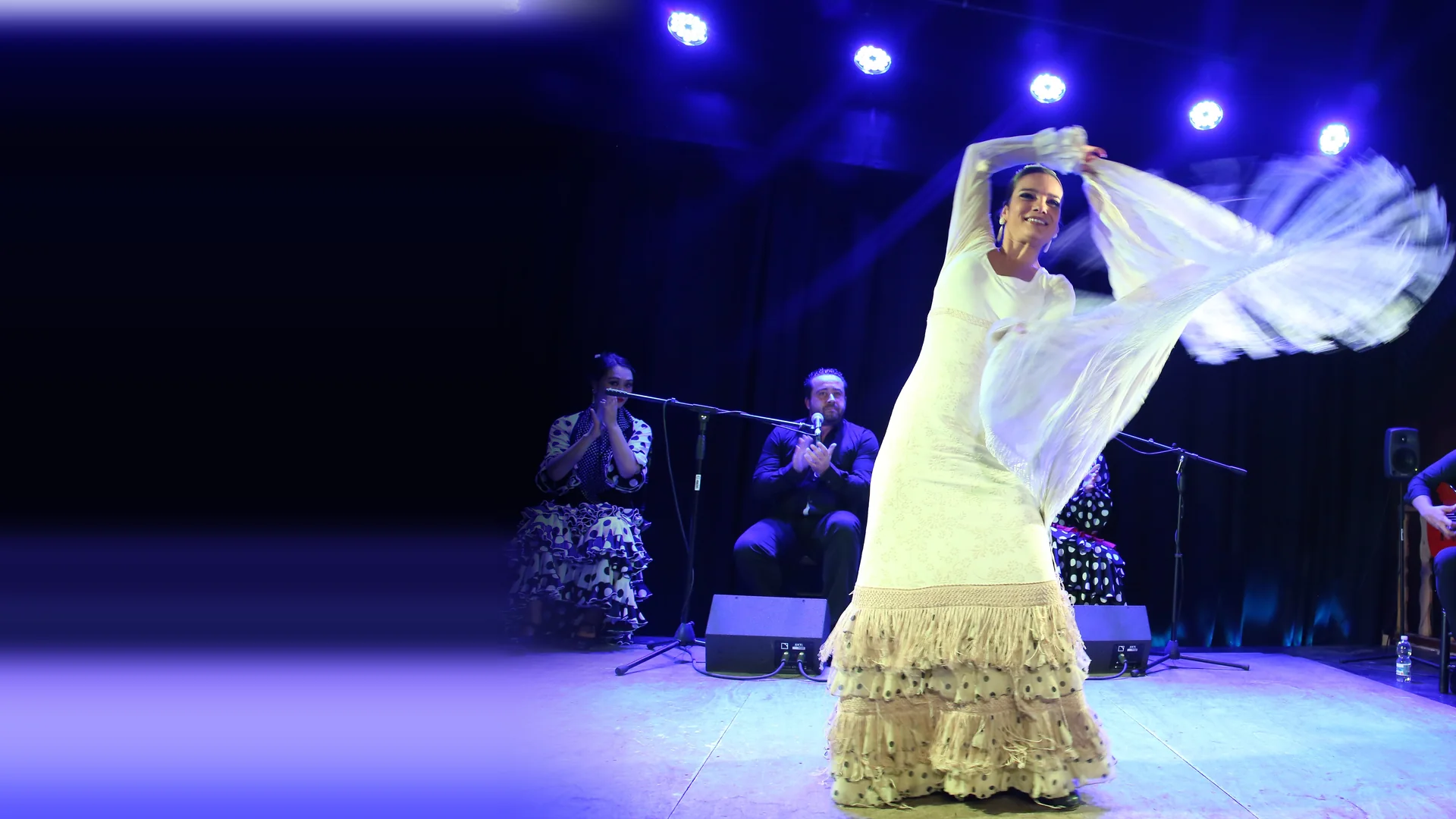 A flamenco dancer in a white dress swirls her shawl on a stage lit in blue, with a singer, a clapping artist and a guitarist seated behind her